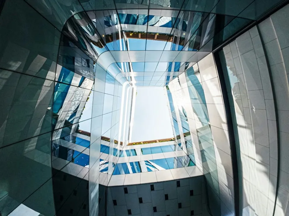 Looking up through a modern glass building interior, with a geometric skylight grid opening to bright sky above.